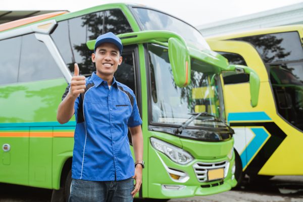 Seorang sopir bus berseragam dan bertopi tersenyum sambil mengacungkan jempol di latar belakang bus